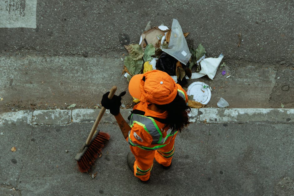 A person dressed in an orange work jumpsuit, wearing white gloves, standing on a concrete floor, holding two large blue plastic rubbish bags, each filled with waste material, with their hands gripping