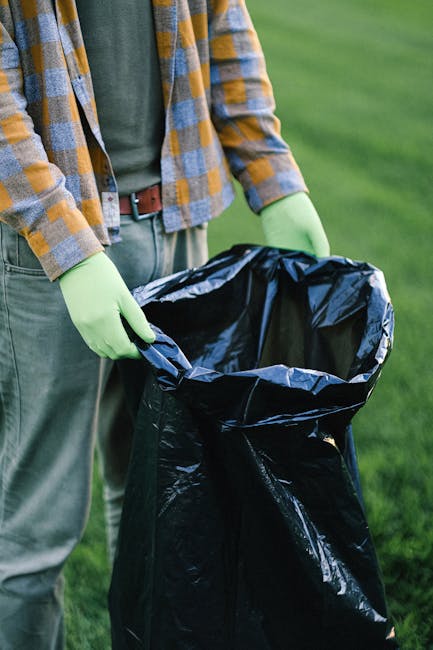 A man operating a backpack leaf blower is standing in a garden or outdoor area with a dirt pathway, using the blower to clear fallen leaves and debris. The man is dressed in work trousers with reflect