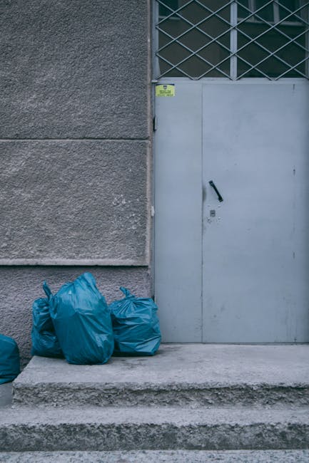 Two waste collection workers are engaged in an outdoor setting during the evening, loading bulky waste into a large, rust-colored metal skip on the back of a waste removal truck. One worker, seen from