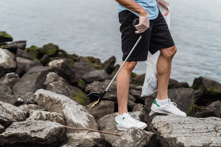 A person wearing a light blue t-shirt, black shorts, and white sneakers is standing on a rocky shoreline near a body of water. They are holding a trash picker in one hand and wearing gloves, while the